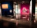 An art gallery with colorful illuminated walls displaying various paintings and artworks. Dimly lit space with modern and abstract art, featuring multiple framed pieces including a large monochrome portrait. Chairs are strategically placed along the walls, and subtle lighting is used to highlight specific artworks.