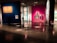 An art gallery with colorful illuminated walls displaying various paintings and artworks. Dimly lit space with modern and abstract art, featuring multiple framed pieces including a large monochrome portrait. Chairs are strategically placed along the walls, and subtle lighting is used to highlight specific artworks.
