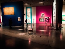 An art gallery with colorful illuminated walls displaying various paintings and artworks. Dimly lit space with modern and abstract art, featuring multiple framed pieces including a large monochrome portrait. Chairs are strategically placed along the walls, and subtle lighting is used to highlight specific artworks.