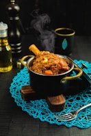 A steaming pot of richly colored cooked food is placed on a decorative blue mat. The pot has a leg of meat with visible bone, surrounded by brown noodles and chunks of vegetables such as carrots. Additional items like a fork, bottle of oil, and a black cup with a gold rim are arranged around the pot.