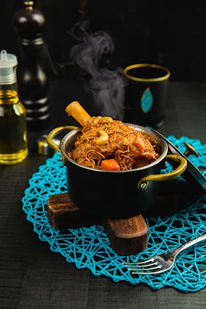 A steaming pot of richly colored cooked food is placed on a decorative blue mat. The pot has a leg of meat with visible bone, surrounded by brown noodles and chunks of vegetables such as carrots. Additional items like a fork, bottle of oil, and a black cup with a gold rim are arranged around the pot.