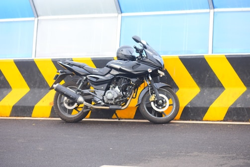 A black motorcycle is parked in front of a barrier with black and yellow chevrons. The background features a structure with blue translucent panels.