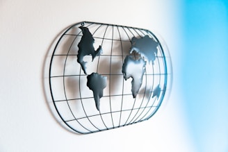 A wall-mounted metal sculpture representing a world map viewed from the side. The continents are formed with metal pieces fixed onto a circular wireframe grid, creating a three-dimensional effect. Soft light casts shadows that enhance the dimensionality of the artwork. A gradient of light blue shades is visible, adding a sense of calmness to the setting.