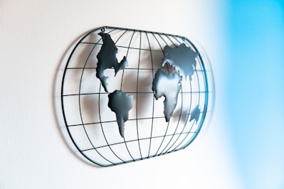 A wall-mounted metal sculpture representing a world map viewed from the side. The continents are formed with metal pieces fixed onto a circular wireframe grid, creating a three-dimensional effect. Soft light casts shadows that enhance the dimensionality of the artwork. A gradient of light blue shades is visible, adding a sense of calmness to the setting.