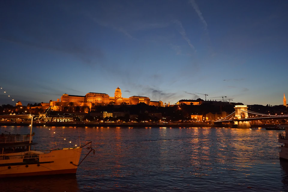 The image depicts a serene night scene at the Buda Castle, located on the banks of the Danube River in Budapest, Hungary. The castle is illuminated by warm orange lights that cast a soft glow over its architectural details and surrounding landscape. In the foreground, two boats are docked along the riverbank, adding to the picturesque setting. The sky above is painted with hues of blue, dotted with wispy clouds, creating an enchanting atmosphere.