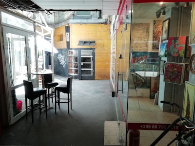 A small indoor space featuring various elements including a glass door with chairs and tables inside, a wooden panelled wall with graffiti, a glass storefront displaying artwork, and a single bike visible in the bottom right corner.
