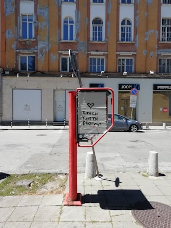 A street scene featuring a red public telephone booth with a message written on its side. The message reads 'Turn on, Tune in, Drop out' with a heart symbol above it. In the background, there is an older, weathered building with a mix of orange and brown facades and arched windows. Several store signs are visible, and a grey car is parked on the street. The pavement is made of square tiles, and small bollards are placed near the booth.
