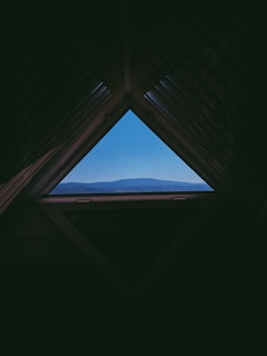 A triangular attic window frames a scenic view of distant blue mountains under a clear sky. The interior of the attic is dark, creating a stark contrast with the bright landscape outside.