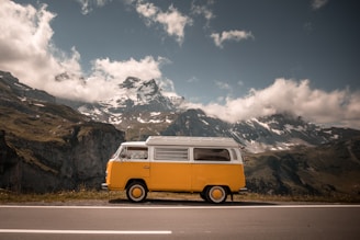 A yellow vintage van is parked by the roadside in a mountainous region with snow-capped peaks and rugged cliffs. The sky is partly cloudy, casting shadows over the landscape.
