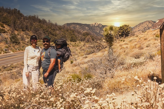 Two adults and a child positioned outdoors in a desert-like landscape. One adult is carrying the child in a backpack suitable for hiking. The vegetation is sparse with dry bushes and a large yucca plant. A roadway runs parallel to the wilderness, and the background features mountain ranges under a setting sun.