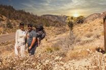 Two adults and a child positioned outdoors in a desert-like landscape. One adult is carrying the child in a backpack suitable for hiking. The vegetation is sparse with dry bushes and a large yucca plant. A roadway runs parallel to the wilderness, and the background features mountain ranges under a setting sun.