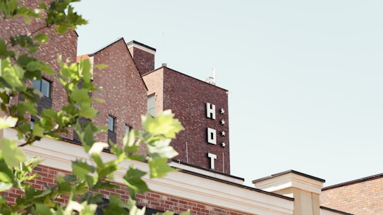 A brick hotel building with a sign that reads 'HOT' and three stars, partially obscured by green leafy branches in the foreground. The sky is clear and light blue, suggesting a bright day.