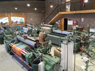 An industrial setup featuring multiple textile machines, likely looms, arranged in rows. The machines are surrounded by spools of thread and fabric in various colors, including a vibrant multicolored textile displayed prominently in the foreground. The room is spacious with brick walls, large windows, and a wooden staircase leading to an upper level. Machinery, piping, and a fire extinguisher are visible, along with safety signs.