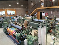 An industrial setup featuring multiple textile machines, likely looms, arranged in rows. The machines are surrounded by spools of thread and fabric in various colors, including a vibrant multicolored textile displayed prominently in the foreground. The room is spacious with brick walls, large windows, and a wooden staircase leading to an upper level. Machinery, piping, and a fire extinguisher are visible, along with safety signs.