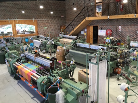 An industrial setup featuring multiple textile machines, likely looms, arranged in rows. The machines are surrounded by spools of thread and fabric in various colors, including a vibrant multicolored textile displayed prominently in the foreground. The room is spacious with brick walls, large windows, and a wooden staircase leading to an upper level. Machinery, piping, and a fire extinguisher are visible, along with safety signs.