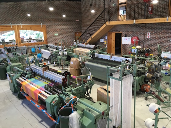 An industrial setup featuring multiple textile machines, likely looms, arranged in rows. The machines are surrounded by spools of thread and fabric in various colors, including a vibrant multicolored textile displayed prominently in the foreground. The room is spacious with brick walls, large windows, and a wooden staircase leading to an upper level. Machinery, piping, and a fire extinguisher are visible, along with safety signs.