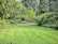 A lush garden with a well-maintained green lawn surrounded by various trees and shrubs. There are colorful flowers and dense foliage creating a serene and vibrant atmosphere. A small, young tree stands in the center, and a rock-bordered area adds texture to the scene.