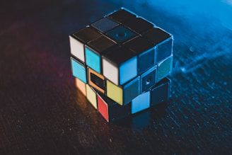 A Rubik's Cube with several colored squares is placed on a dark wooden surface. The lighting casts a blue hue over part of the image, creating a contrast between the cube's vibrant colors and the surrounding darkness.