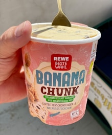 A hand is holding a container of 'Banana Chunk' vegan ice cream by Rewe Beste Wahl. The ice cream features flavors of dark chocolate and walnut pieces. A spoon is inserted into the creamy, light-colored dessert.