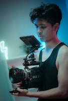A person holding a professional camera setup with a focus on the equipment. The camera has a large lens and an attached monitor. The lighting appears moody with hues of blue and red, enhancing the cinematic feel.