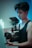A person holding a professional camera setup with a focus on the equipment. The camera has a large lens and an attached monitor. The lighting appears moody with hues of blue and red, enhancing the cinematic feel.