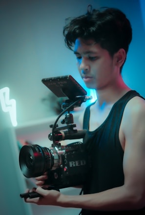 A person holding a professional camera setup with a focus on the equipment. The camera has a large lens and an attached monitor. The lighting appears moody with hues of blue and red, enhancing the cinematic feel.
