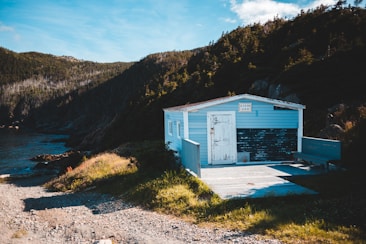 A small, weathered blue cabin with a sign that reads 'Ocean View' is situated on a rocky coastline. The cabin has peeling paint and an adjacent wooden deck with a bench. Surrounding the cabin are lush green grasses and tall, forested hills. The ocean is visible to the left, with gentle waves and a clear sky overhead.