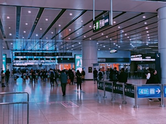 A busy indoor scene of a transit station with several people walking and standing around. Overhead signs display directions and information in both Chinese and English. The flooring is shiny and reflective, while the ceiling features bright lighting panels. Some shops with colorful signage are visible in the background.