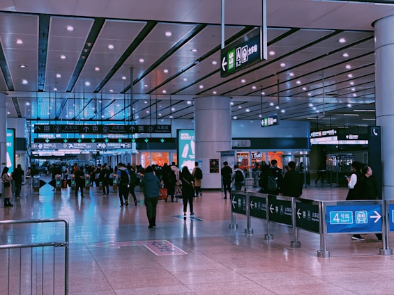 A busy indoor scene of a transit station with several people walking and standing around. Overhead signs display directions and information in both Chinese and English. The flooring is shiny and reflective, while the ceiling features bright lighting panels. Some shops with colorful signage are visible in the background.