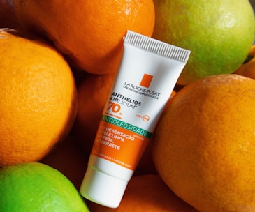 A tube of La Roche-Posay Anthelios sunscreen is placed among a group of oranges and a lime. The oranges have a bright orange hue, and the lime is green. The sunscreen tube features orange, green, and white colors, and displays SPF 70 prominently.