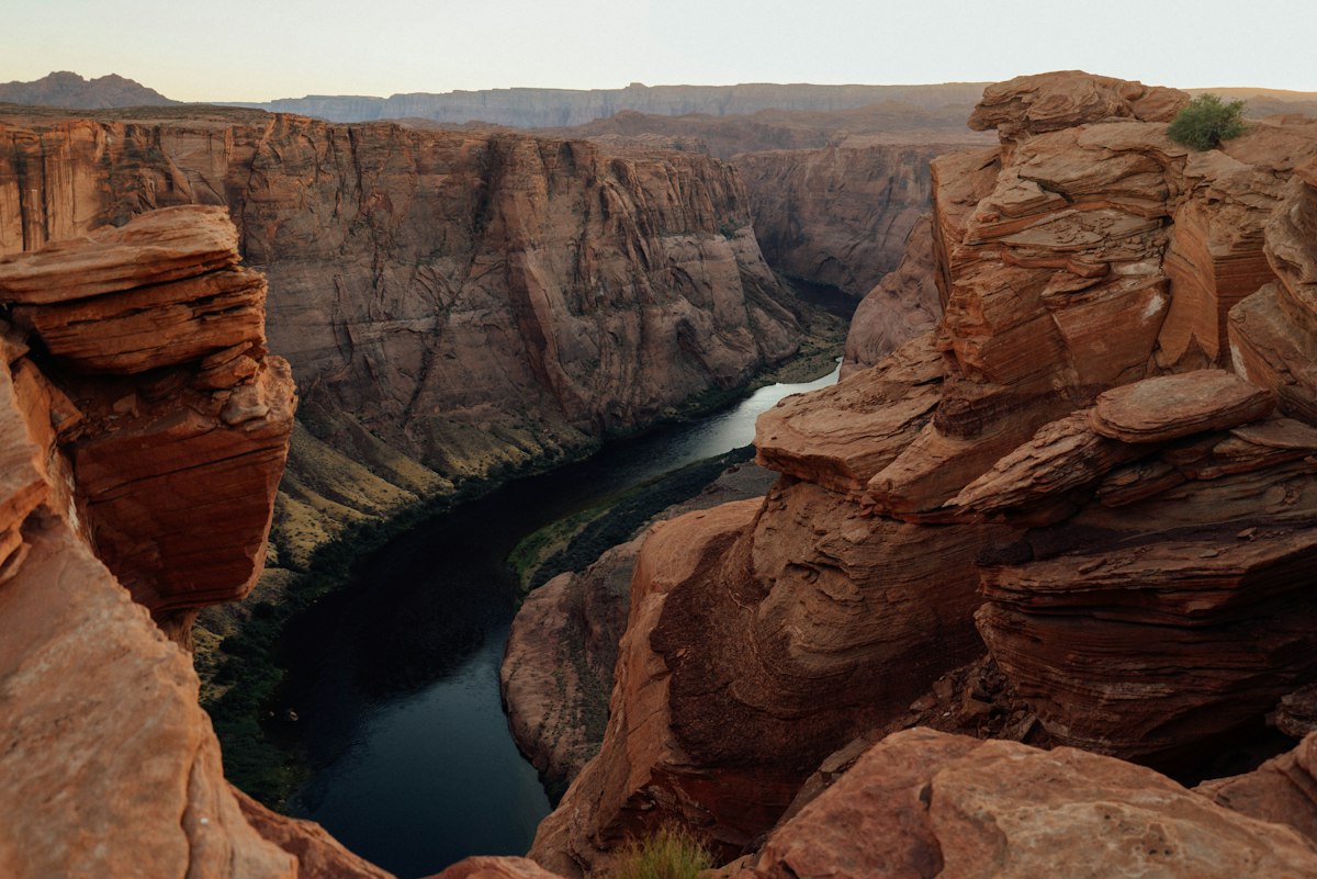 The Colorado River winding between layered red sandstone canyon walls seen from above, bleached rock ledges in the foreground, dark water below and a hazy ridge on the horizon