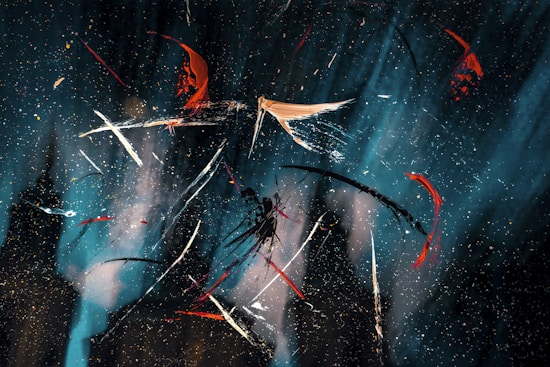 Abstract art featuring dynamic streaks of paint in various colors including white, red, orange, and black on a dark blue and black background with scattered speckles. The strokes create a sense of movement and chaos, with the colors contrasting sharply against the darker hues.