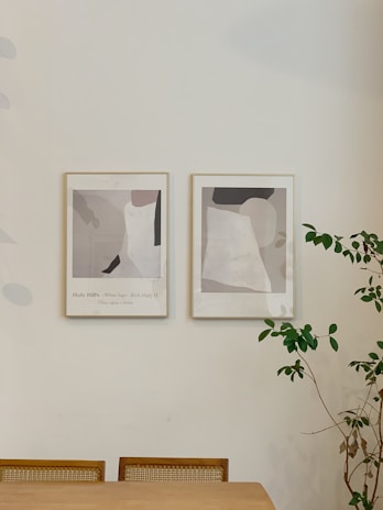 Two framed abstract art prints are hung on a light-colored wall above a wooden table with simple cane-back chairs. To the right, a leafy green plant adds a touch of nature to the scene.