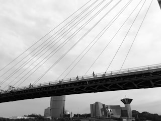 A black and white image of a large bridge with cables extending diagonally across. Several people can be seen walking along the bridge. Below the bridge, modern architectural structures with unique shapes are visible, including cylindrical and angular buildings.