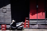 A white scooter is parked in front of a wooden building with slatted walls, featuring a combination of dark and red panels. Several red plastic crates with labels are stacked and placed next to the scooter, casting shadows on the ground.