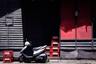 A white scooter is parked in front of a wooden building with slatted walls, featuring a combination of dark and red panels. Several red plastic crates with labels are stacked and placed next to the scooter, casting shadows on the ground.