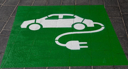 A green painted square on a pavement contains a white stencil of a car with a cable and plug, symbolizing an electric vehicle charging station.