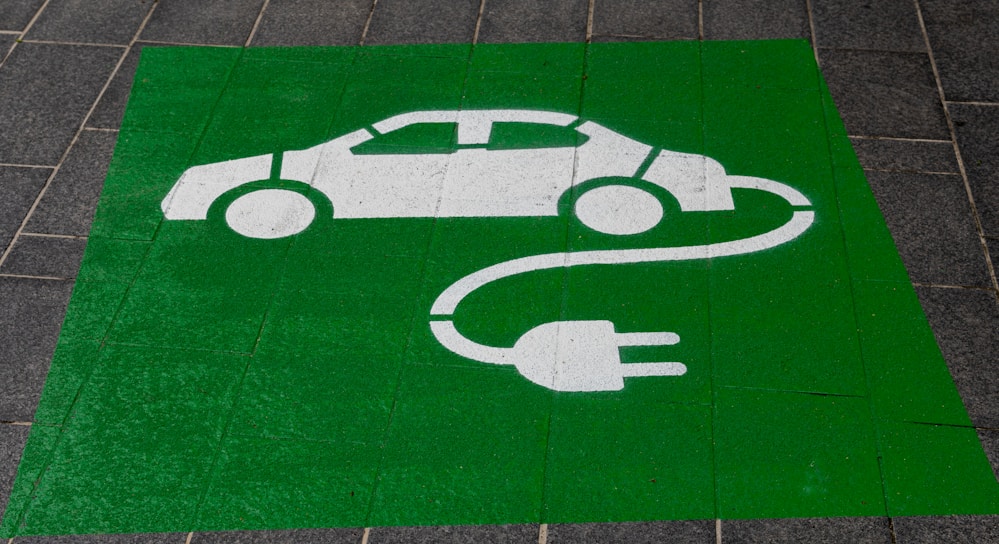 A green painted square on a pavement contains a white stencil of a car with a cable and plug, symbolizing an electric vehicle charging station.