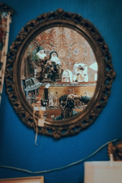 A decorative oval mirror with an intricate frame reflects a scene that includes a person taking a photo with a camera and various decorative items such as Santa Claus figurines and other collectibles arranged on a shelf. The background features a patterned tapestry, and the overall setting appears to be a cozy, eclectic room.