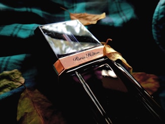 A perfume bottle with reflective surfaces and the label 'Paris Hilton' is surrounded by dried leaves. The bottle has a metallic and glossy finish, placed on a dark green checkered fabric that adds a rich texture to the composition.