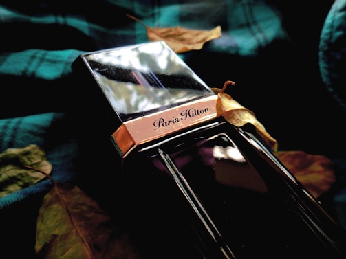 A perfume bottle with reflective surfaces and the label 'Paris Hilton' is surrounded by dried leaves. The bottle has a metallic and glossy finish, placed on a dark green checkered fabric that adds a rich texture to the composition.