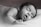A black and white photo of a baby peacefully sleeping on a soft surface with eyes closed and mouth slightly open. The infant's head rests gently on one arm, and the atmosphere is calm and serene.