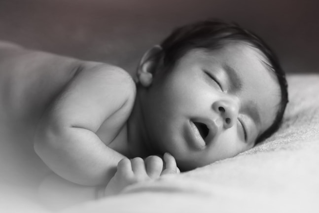 A black and white photo of a baby peacefully sleeping on a soft surface with eyes closed and mouth slightly open. The infant's head rests gently on one arm, and the atmosphere is calm and serene.