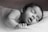 A black and white photo of a baby peacefully sleeping on a soft surface with eyes closed and mouth slightly open. The infant's head rests gently on one arm, and the atmosphere is calm and serene.