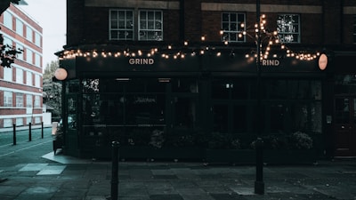 A street corner features a dark-colored cafe with glowing string lights hanging above the entrance. The sign 'Grind' is prominently displayed on the facade. The surrounding area includes a paved sidewalk and a brick building across the street, with some trees partially visible.