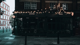 A street corner features a dark-colored cafe with glowing string lights hanging above the entrance. The sign 'Grind' is prominently displayed on the facade. The surrounding area includes a paved sidewalk and a brick building across the street, with some trees partially visible.