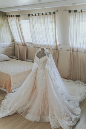 A detailed and elegant wedding dress on a mannequin in a softly lit bedroom. The dress features intricate lace detailing and a long, flowing train. Light streams in through sheer, pleated curtains that frame the windows, creating a serene and romantic atmosphere. The room includes a neatly made bed with a neutral-colored bedspread, enhancing the calm and delicate setting.