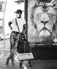 A person wearing a mask and a cap leans against a billboard featuring a lion's face. The individual has a shopping cart with bags, standing casually on a paved sidewalk. The lion on the billboard appears calm and majestic, with its eyes closed.