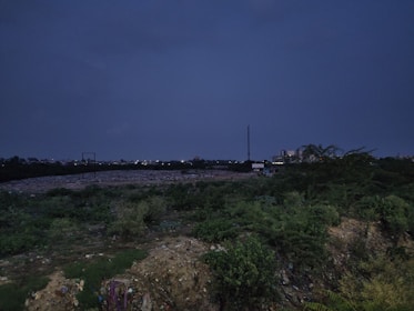 A dark, expansive landscape with dense vegetation in the foreground and a large area that appears to be a landfill or wasteland in the midground. There are small structures or machinery, possibly for waste management, scattered around the area. In the distance, city buildings and lights are visible below the dark, overcast sky.