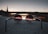 A pair of round sunglasses with tinted lenses rests on a concrete surface, with a blurred view of a bridge and urban skyline in the background. The scene is captured during a sunset, creating a gradient of warm and cool hues in the sky.